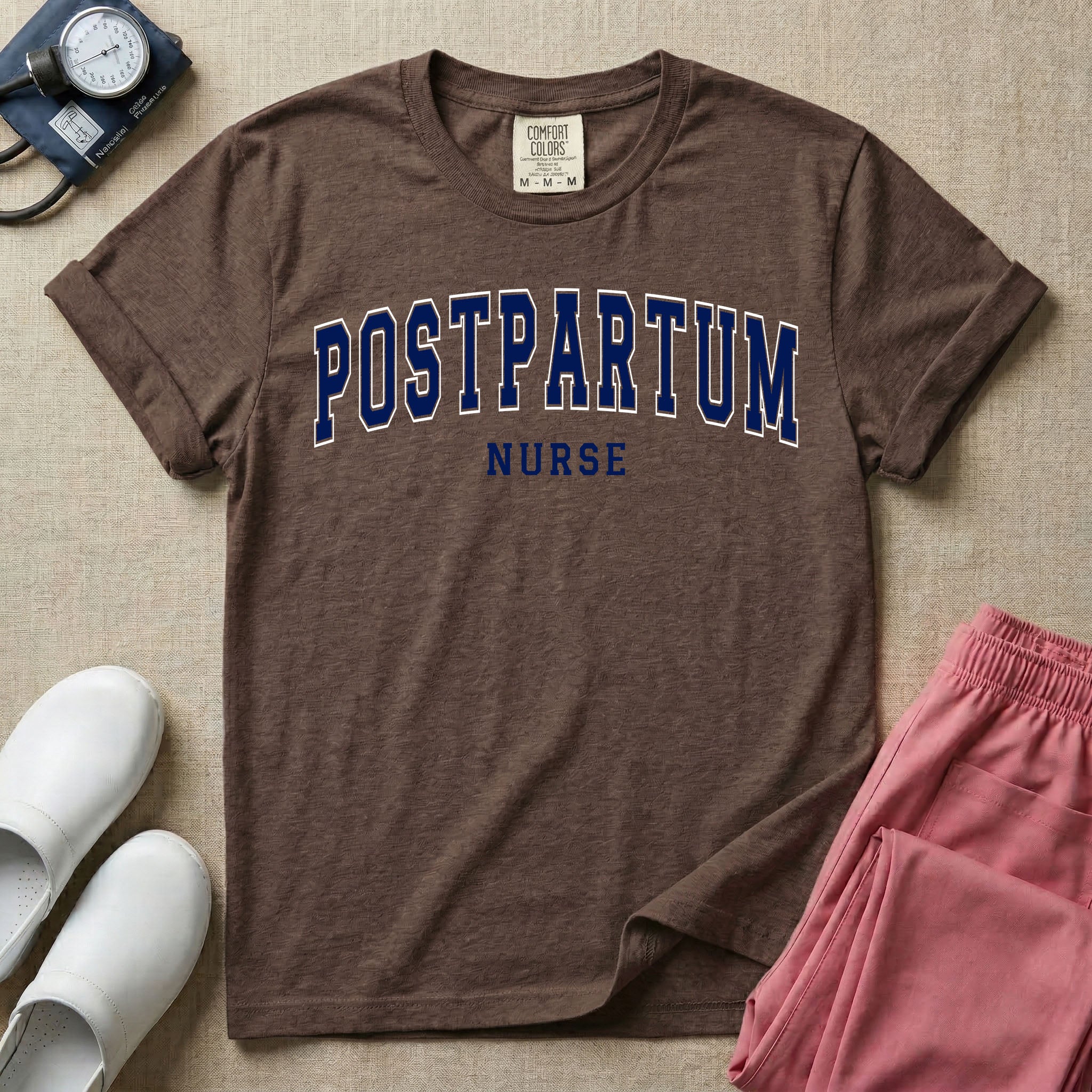 A t-shirt featuring the word POSTPARTUM arched in large collegiate block letters above the word NURSE in a smaller, matching font.