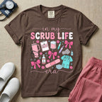 A cute general nursing t-shirt featuring the retro text "in my SCRUB LIFE era" surrounded by coquette medical graphics including a pink tumbler cup, stethoscope, IV bag with pink bows, clipboard, and blue scrubs.