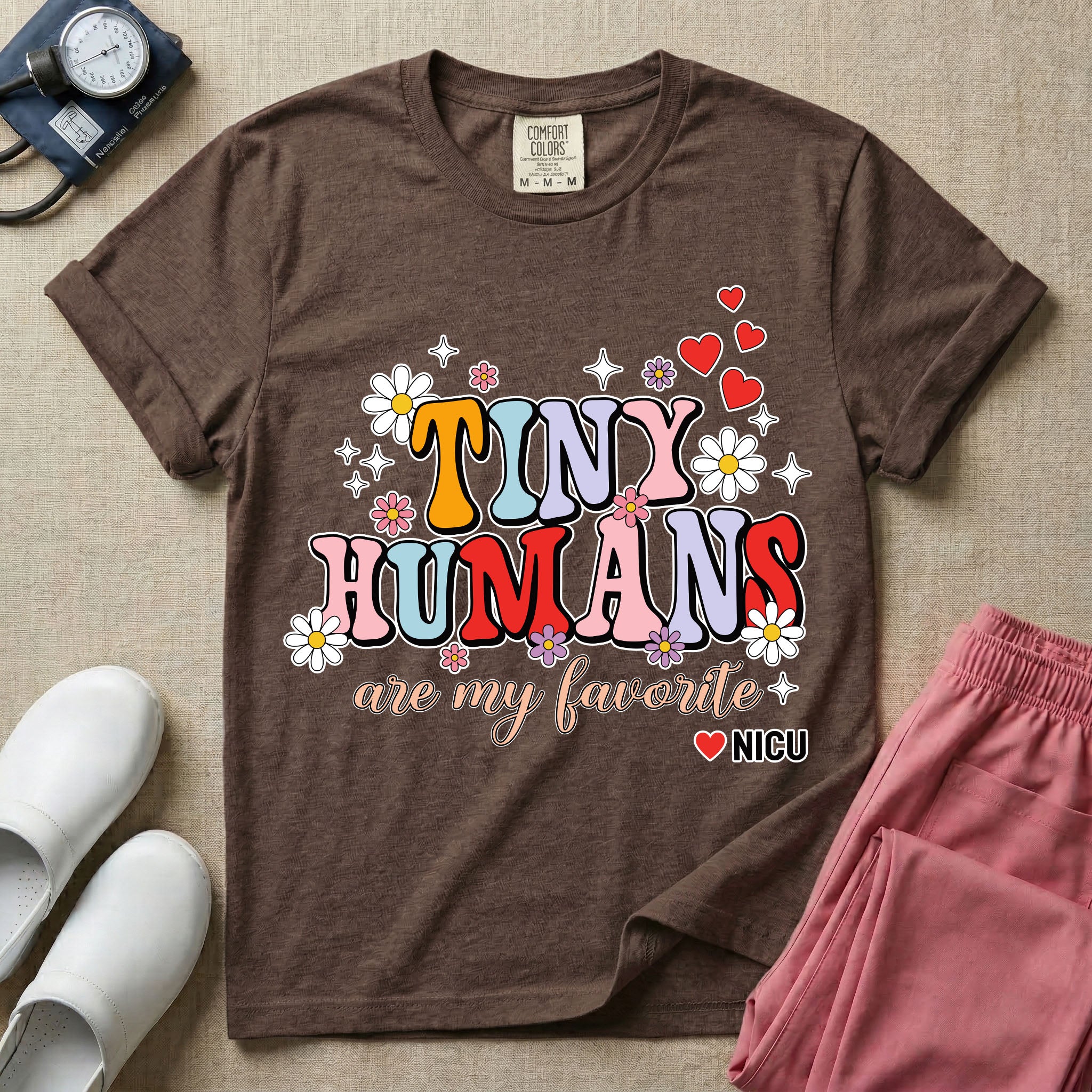 A t-shirt featuring the text Tiny Humans are my favorite in colorful retro bubble letters decorated with daisies, sparkles, and red hearts with a small NICU heart logo at the bottom.