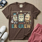 A retro-style t-shirt for emergency room nurses featuring the text "ER CREW" below a vintage cartoon illustration of a medicine bottle, a grumpy clipboard holding an AMA form, and a sandwich wearing a nurse cap walking together.