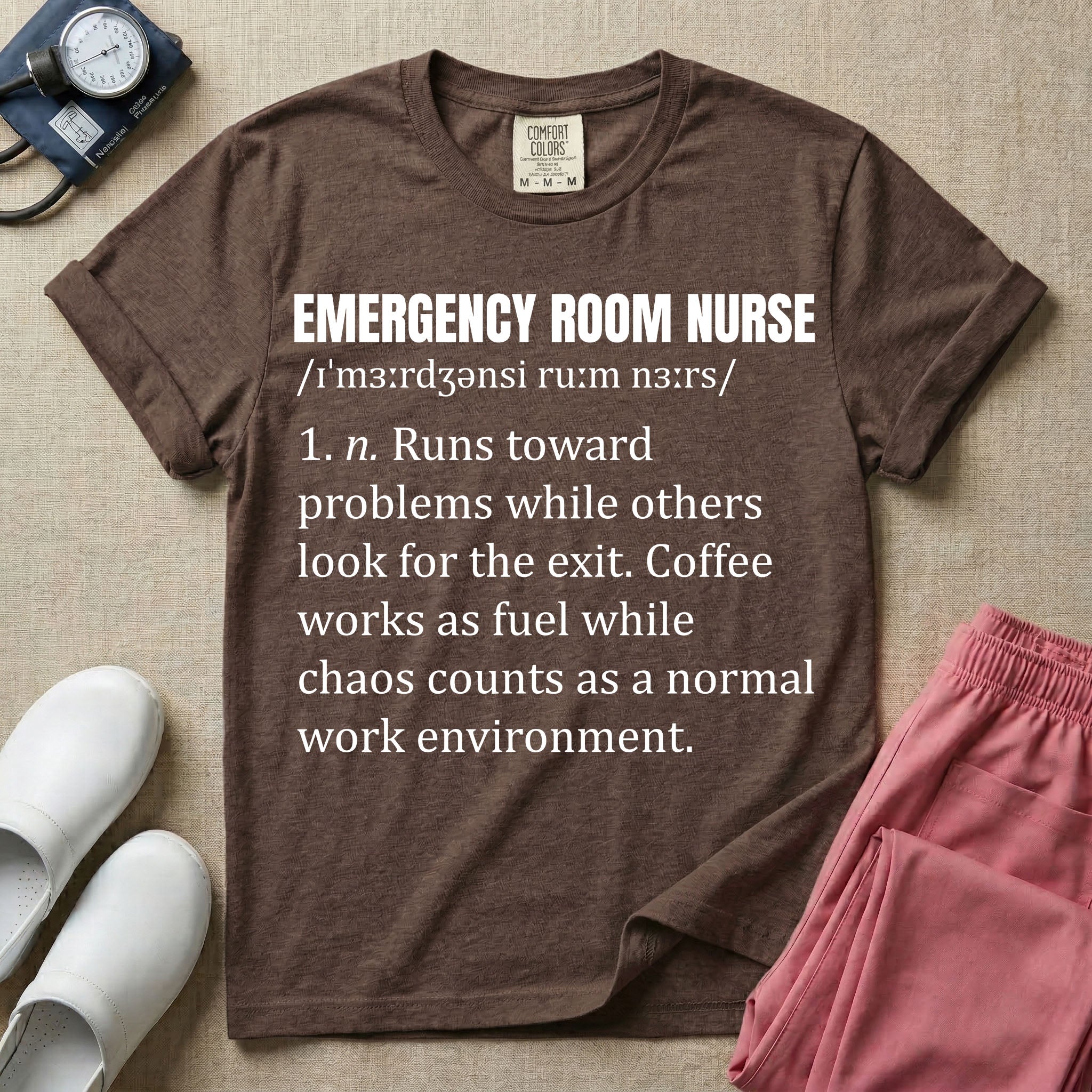 A minimalist t-shirt for emergency room nurses featuring a dictionary definition graphic for the term "EMERGENCY ROOM NURSE" with a phonetic pronunciation and a humorous description about running toward problems, using coffee as fuel, and thriving in chaos.