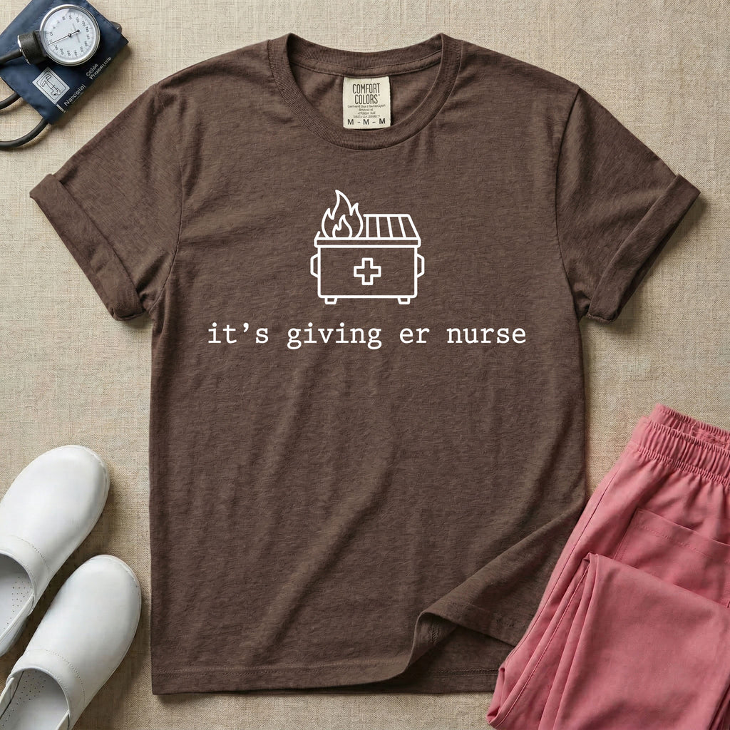A funny minimalist ER nursing t-shirt featuring a simple white line drawing of a dumpster on fire with a medical cross on it, sitting above the lower-case typewriter text "it's giving er nurse".