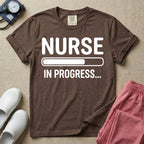 General nursing student t-shirt featuring a white loading bar graphic and the text "NURSE IN PROGRESS..." on a dark gray garment, in espresso color.