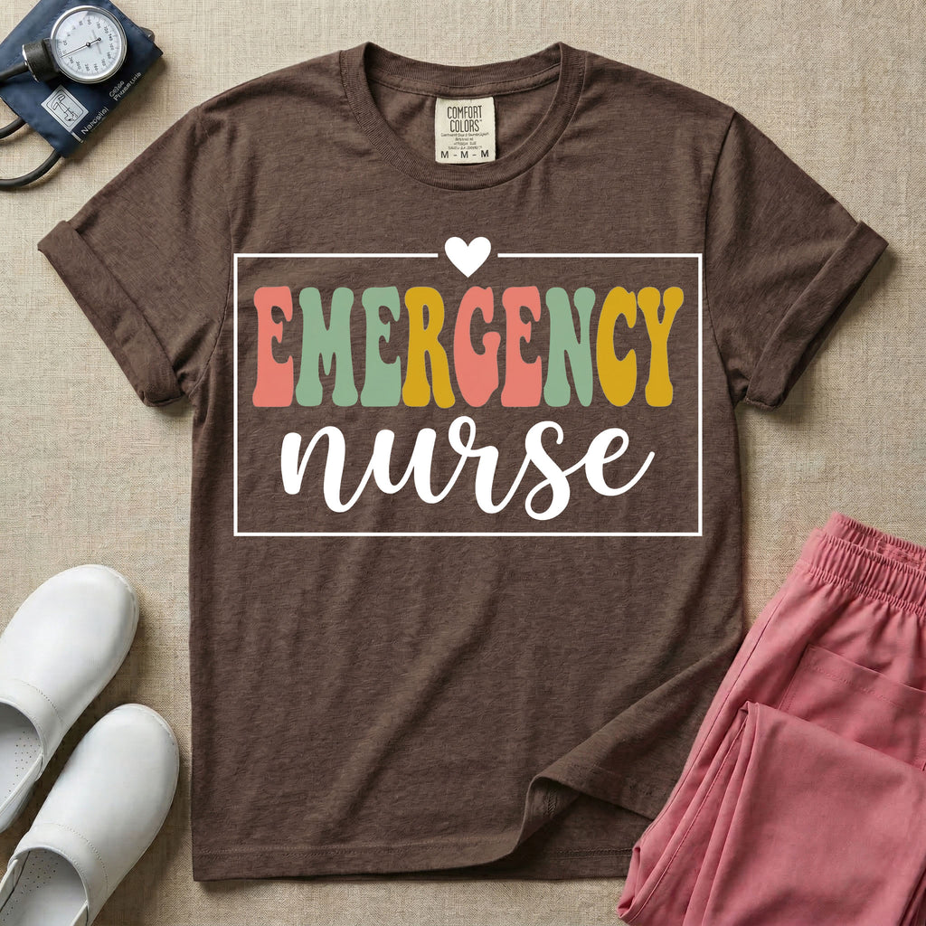 A retro-style t-shirt for emergency room nurses featuring the word "EMERGENCY" in colorful, vintage lettering above the word "nurse" in a delicate white script font, framed by a thin white rectangular border with a small heart at the top center.