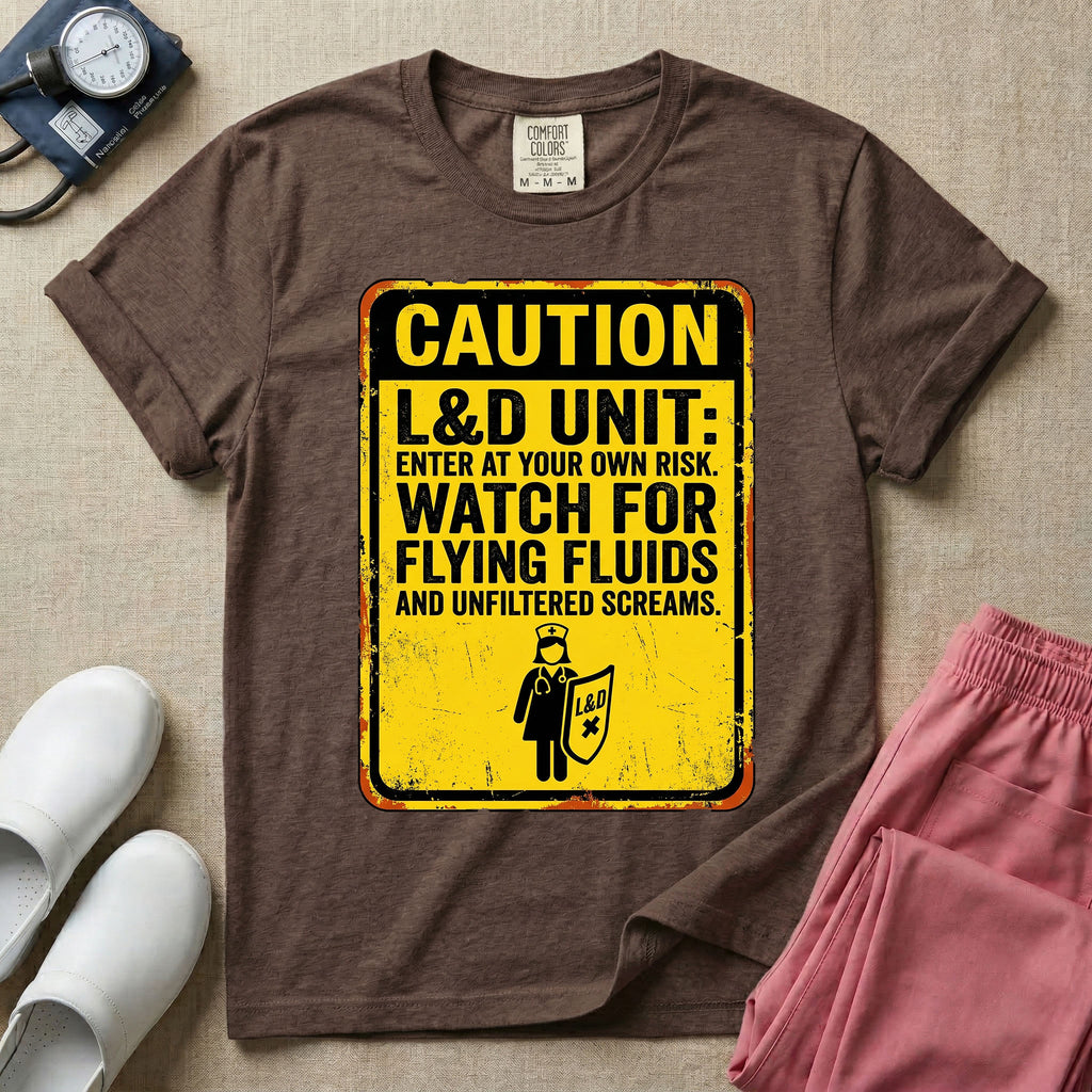 A humorous graphic tee for labor and delivery nurses featuring a distressed caution sign design with the text "CAUTION L&D UNIT: ENTER AT YOUR OWN RISK. WATCH FOR FLYING FLUIDS AND UNFILTERED SCREAMS." above a stick figure nurse holding a shield.