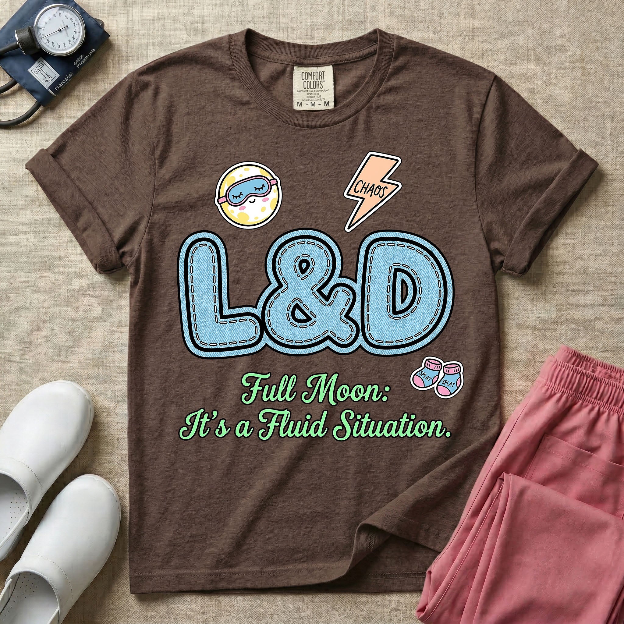 A humorous Labor and Delivery nurse graphic featuring large, light blue varsity-style letters spelling L&D with a pink dashed outline, surrounded by small patch-like icons of a baby and a lightning bolt, sitting above cursive text that reads Full Moon: It's a Fluid Situation.