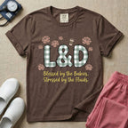 A descriptive graphic tee for labor and delivery nurses featuring plaid block letters spelling L&D surrounded by floral elements and a coffee cup, with the text "Blessed by the Babies, Stressed by the Fluids."