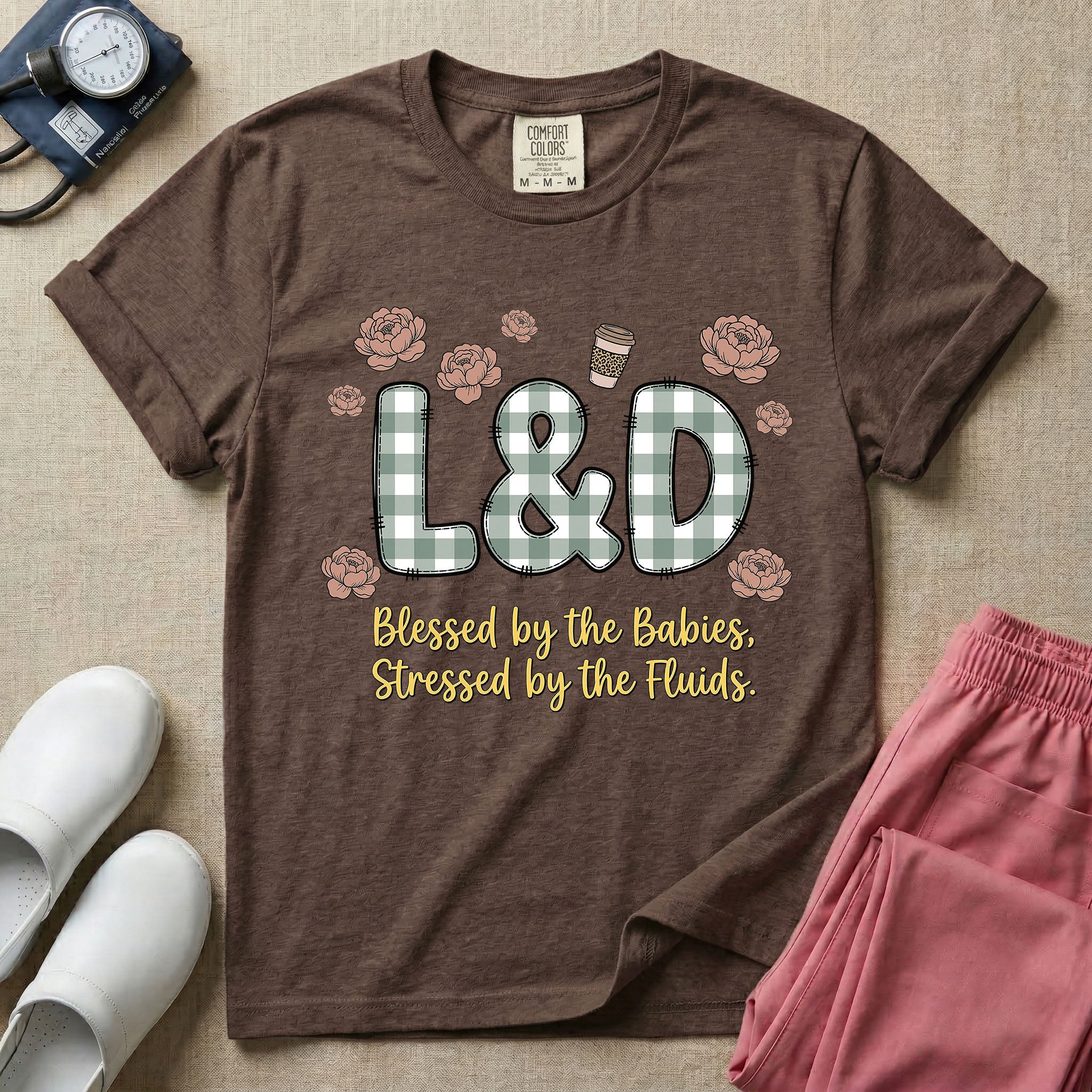 A descriptive graphic tee for labor and delivery nurses featuring plaid block letters spelling L&D surrounded by floral elements and a coffee cup, with the text "Blessed by the Babies, Stressed by the Fluids."