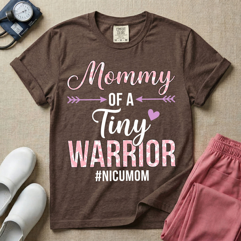 A cute NICU mom t-shirt featuring the stacked text "Mommy OF A Tiny WARRIOR #NICUMOM" in pink and white, accented with decorative arrows, a small pink heart, and a pink plaid pattern filling the word warrior.