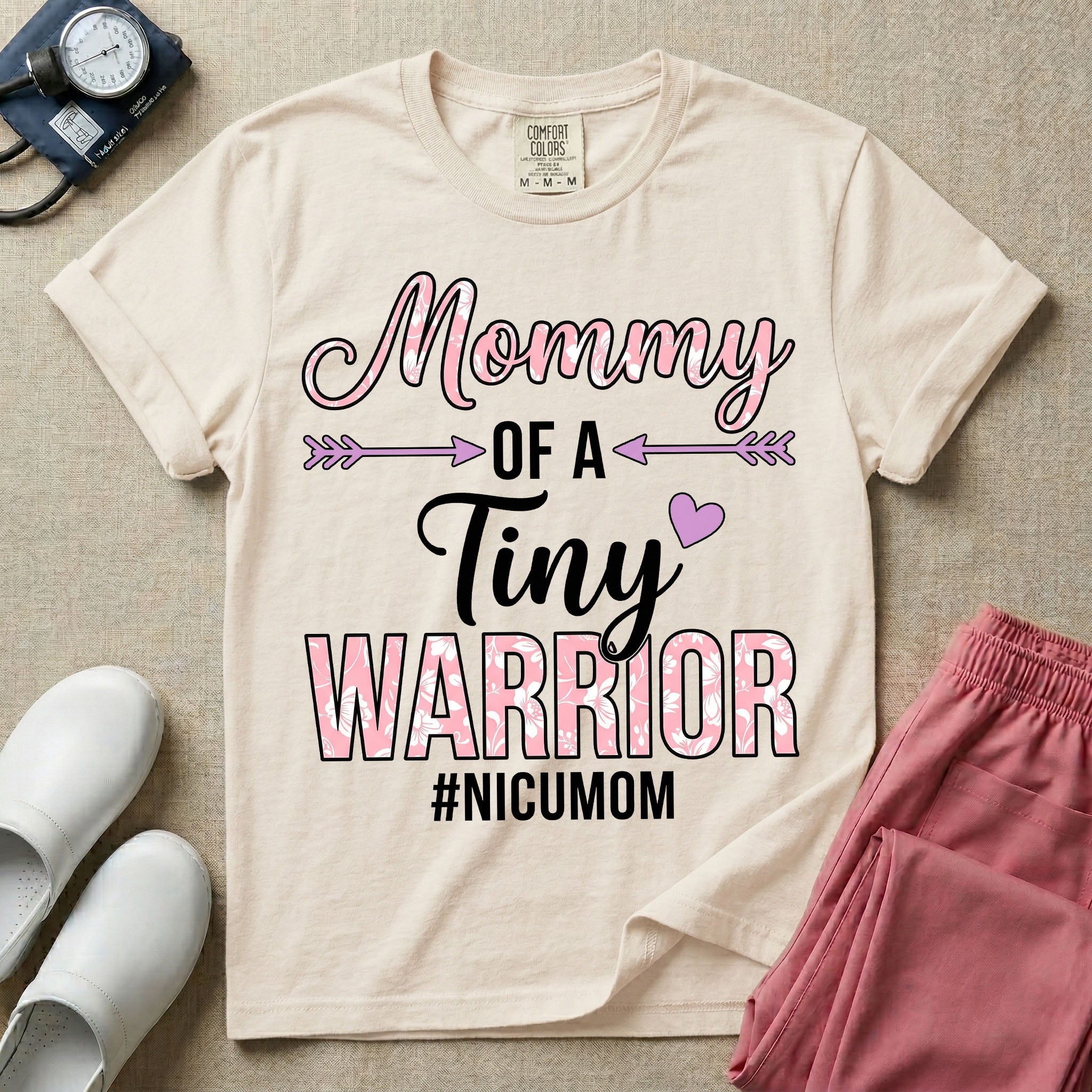 A cute NICU mom t-shirt featuring the stacked text "Mommy OF A Tiny WARRIOR #NICUMOM" in pink and white, accented with decorative arrows, a small pink heart, and a pink plaid pattern filling the word warrior.