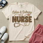 A graphic t-shirt for a Labor and Delivery unit featuring the text 'Labor & Delivery' in beige script above the word 'NURSE' in large leopard print letters, intertwined with an illustration of a stethoscope that features a small red heart on the chest piece.