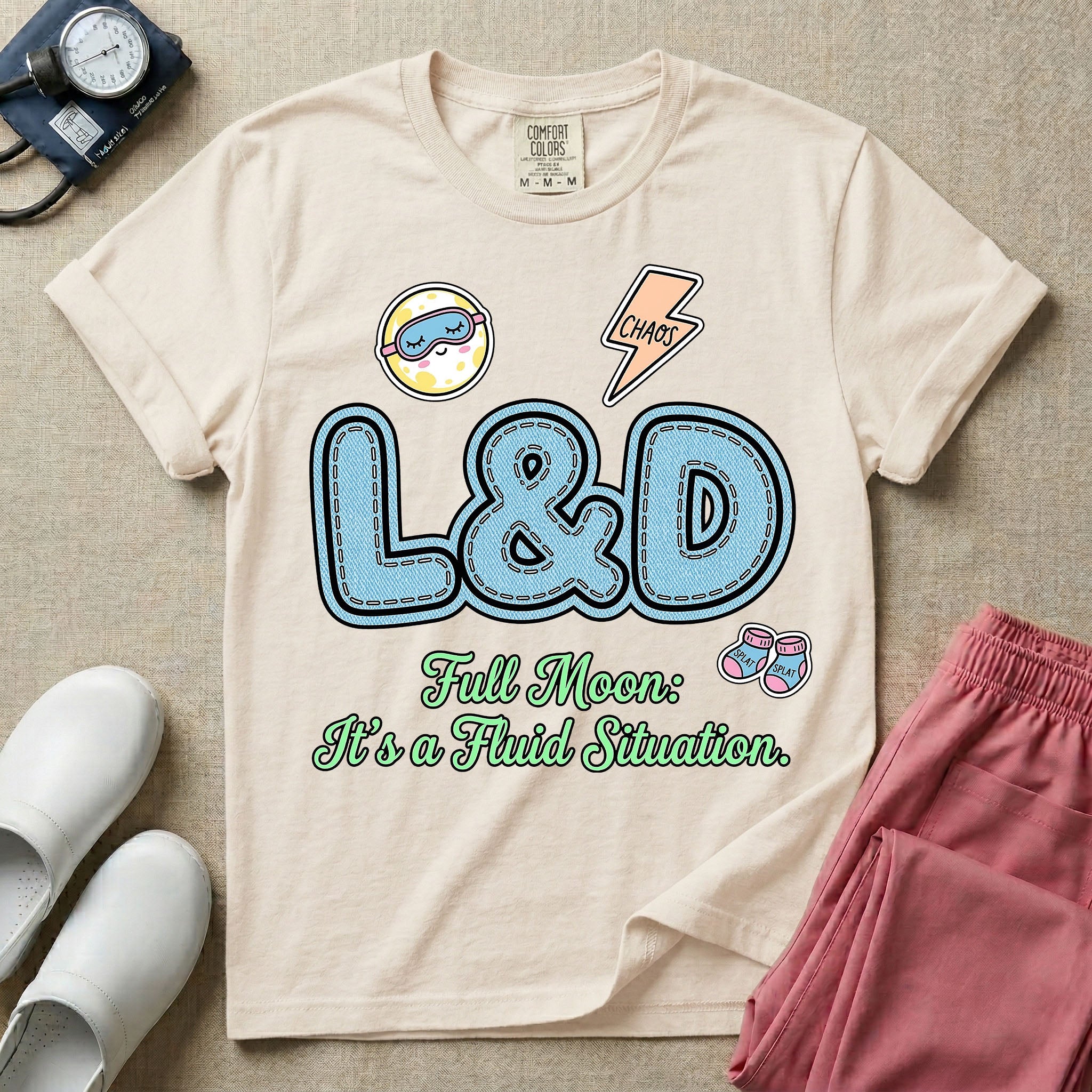 A humorous Labor and Delivery nurse graphic featuring large, light blue varsity-style letters spelling L&D with a pink dashed outline, surrounded by small patch-like icons of a baby and a lightning bolt, sitting above cursive text that reads Full Moon: It's a Fluid Situation.