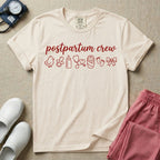 A t-shirt featuring the lowercase text postpartum crew in a cursive script font above a horizontal row of minimalist line-art icons including a baby chick, a pacifier, a baby bottle, a heart-shaped stethoscope, a swaddled baby, baby footprints, and a ribbon bow.
