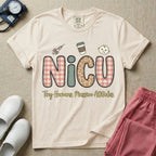 A humorous t-shirt for NICU nurses featuring the large text "NICU" in plaid and patterned letters with the phrase "Tiny Humans. Massive Attitudes." underneath, accompanied by small cute graphics of a syringe, an iced coffee cup, and a baby's face.