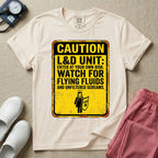A humorous graphic tee for labor and delivery nurses featuring a distressed caution sign design with the text "CAUTION L&D UNIT: ENTER AT YOUR OWN RISK. WATCH FOR FLYING FLUIDS AND UNFILTERED SCREAMS." above a stick figure nurse holding a shield.
