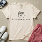A funny minimalist ER nursing t-shirt featuring a simple white line drawing of a dumpster on fire with a medical cross on it, sitting above the lower-case typewriter text "it's giving er nurse".