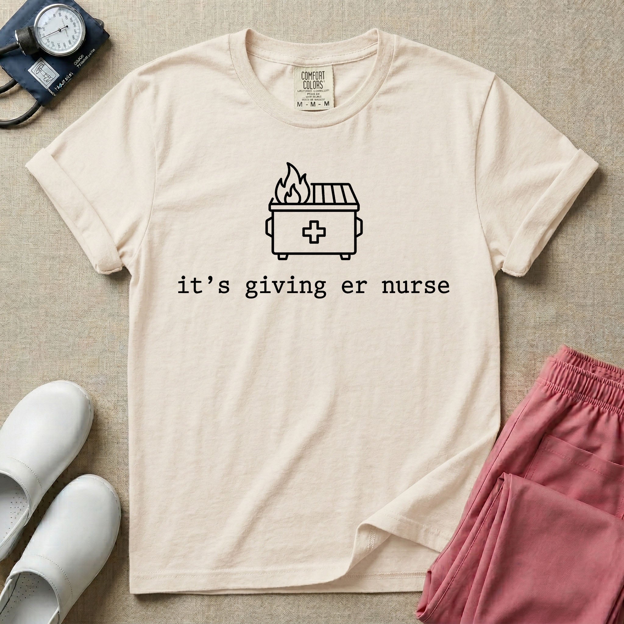 A funny minimalist ER nursing t-shirt featuring a simple white line drawing of a dumpster on fire with a medical cross on it, sitting above the lower-case typewriter text "it's giving er nurse".