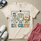A retro-style t-shirt for emergency room nurses featuring the text "ER CREW" below a vintage cartoon illustration of a medicine bottle, a grumpy clipboard holding an AMA form, and a sandwich wearing a nurse cap walking together.
