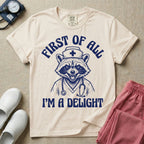 A humorous graphic of a raccoon dressed in a nursing cap and scrub jacket with a stethoscope, with the text "FIRST OF ALL I'M A DELIGHT", in ivory color.