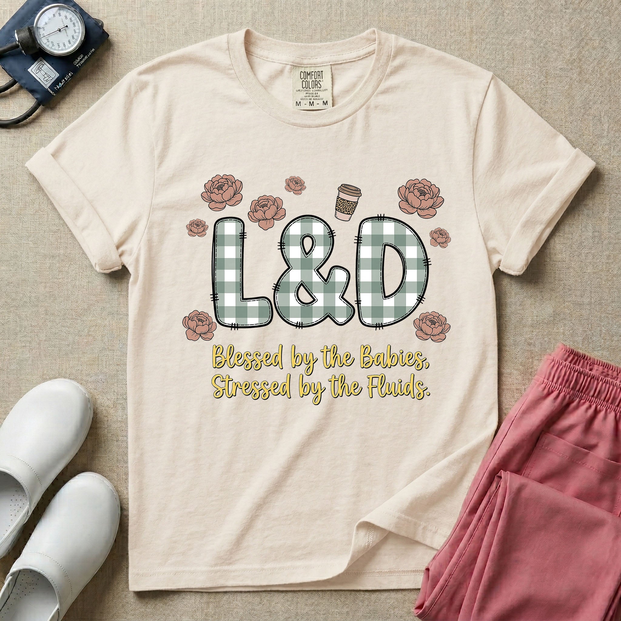 A descriptive graphic tee for labor and delivery nurses featuring plaid block letters spelling L&D surrounded by floral elements and a coffee cup, with the text "Blessed by the Babies, Stressed by the Fluids."