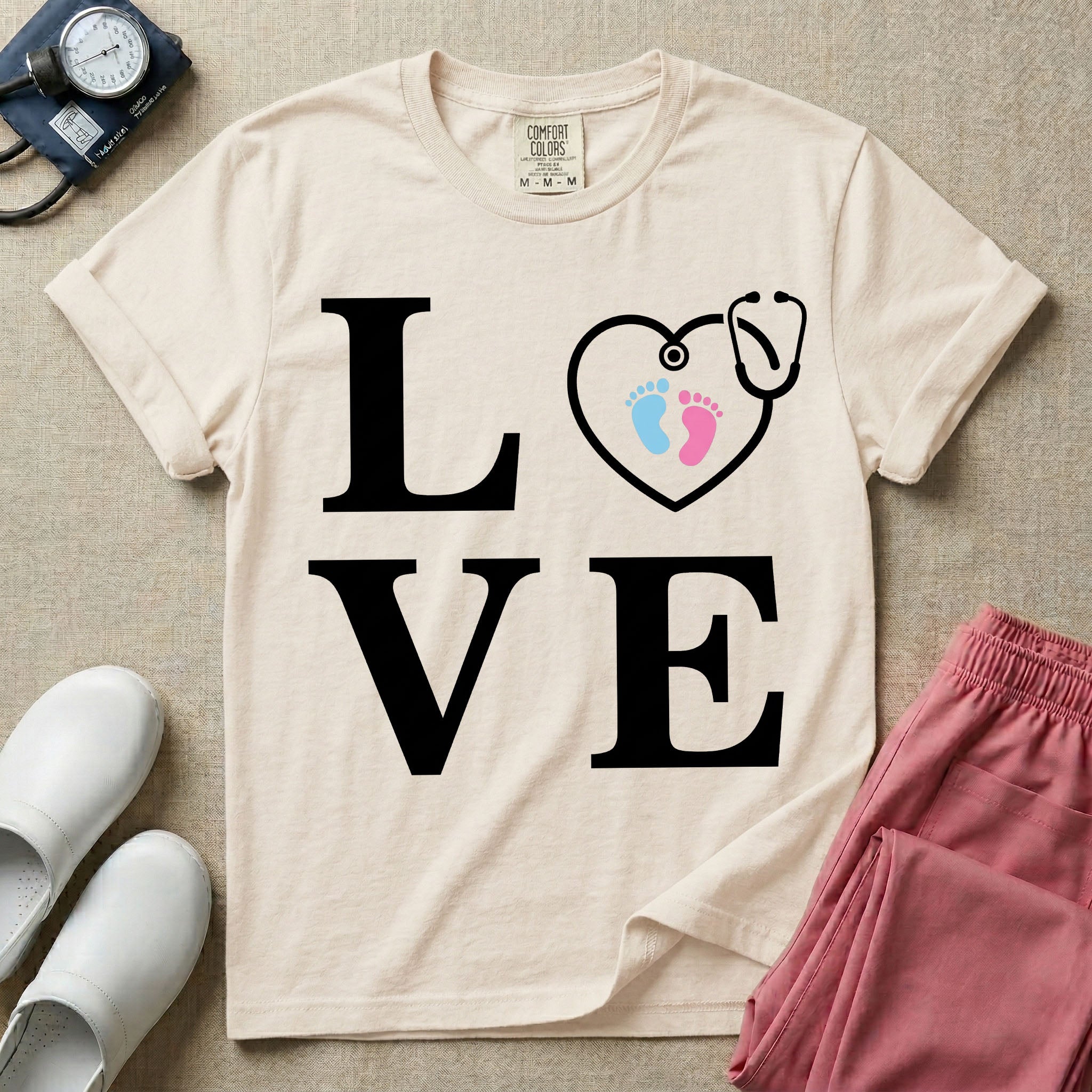 A minimalist nursing t-shirt featuring the word LOVE stacked in large white serif letters, with the letter O replaced by a white stethoscope shaped like a heart containing two small baby footprints, one pink and one blue.