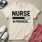 General nursing student t-shirt featuring a white loading bar graphic and the text "NURSE IN PROGRESS..." on a dark gray garment, in ivory color.