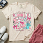 A cute general nursing t-shirt featuring the retro text "in my SCRUB LIFE era" surrounded by coquette medical graphics including a pink tumbler cup, stethoscope, IV bag with pink bows, clipboard, and blue scrubs.