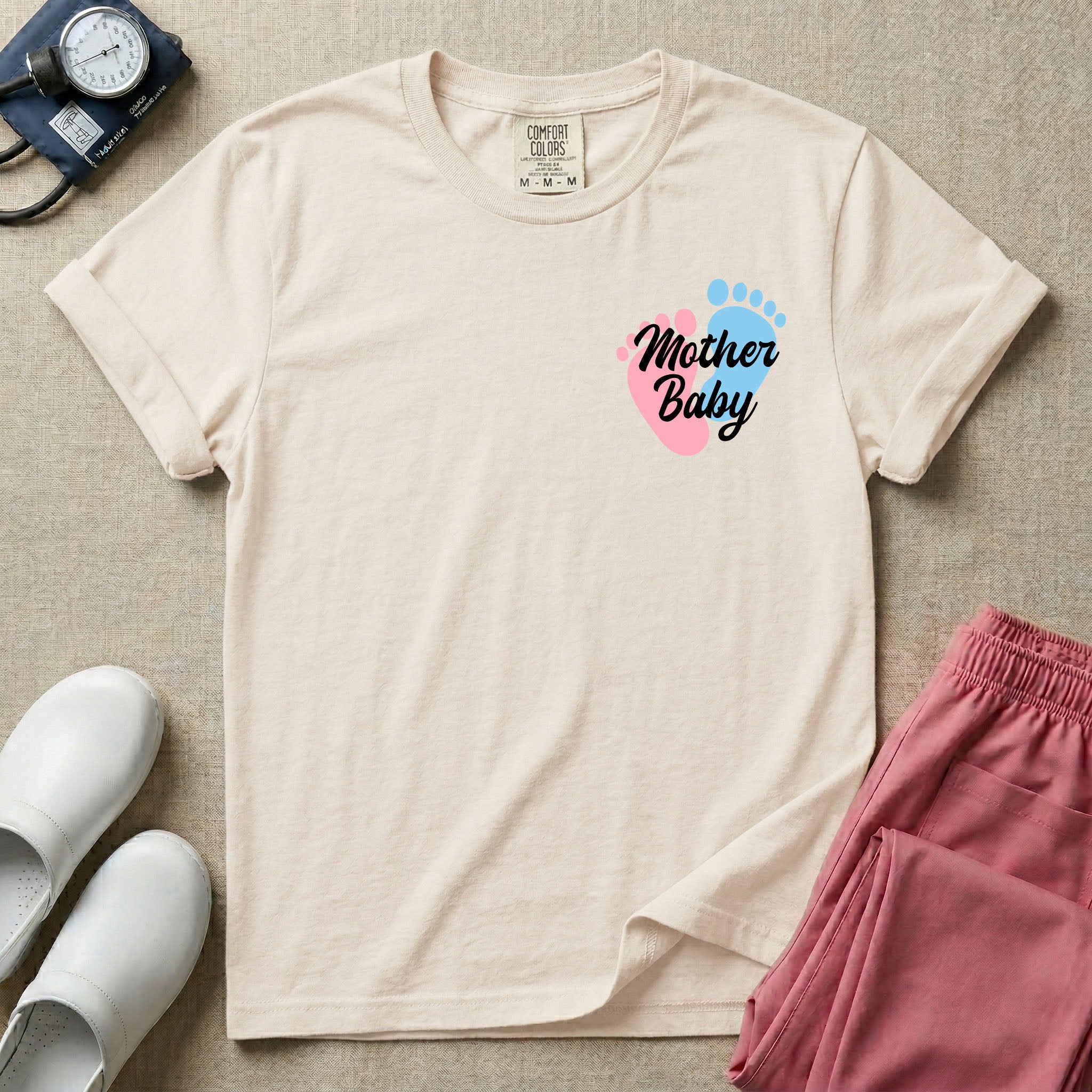 A minimalist postpartum nursing t-shirt featuring a small left chest graphic of two baby footprints, one pink and one blue, with the cursive white text "Mother Baby" written across them.