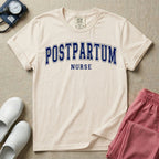 A t-shirt featuring the word POSTPARTUM arched in large collegiate block letters above the word NURSE in a smaller, matching font.