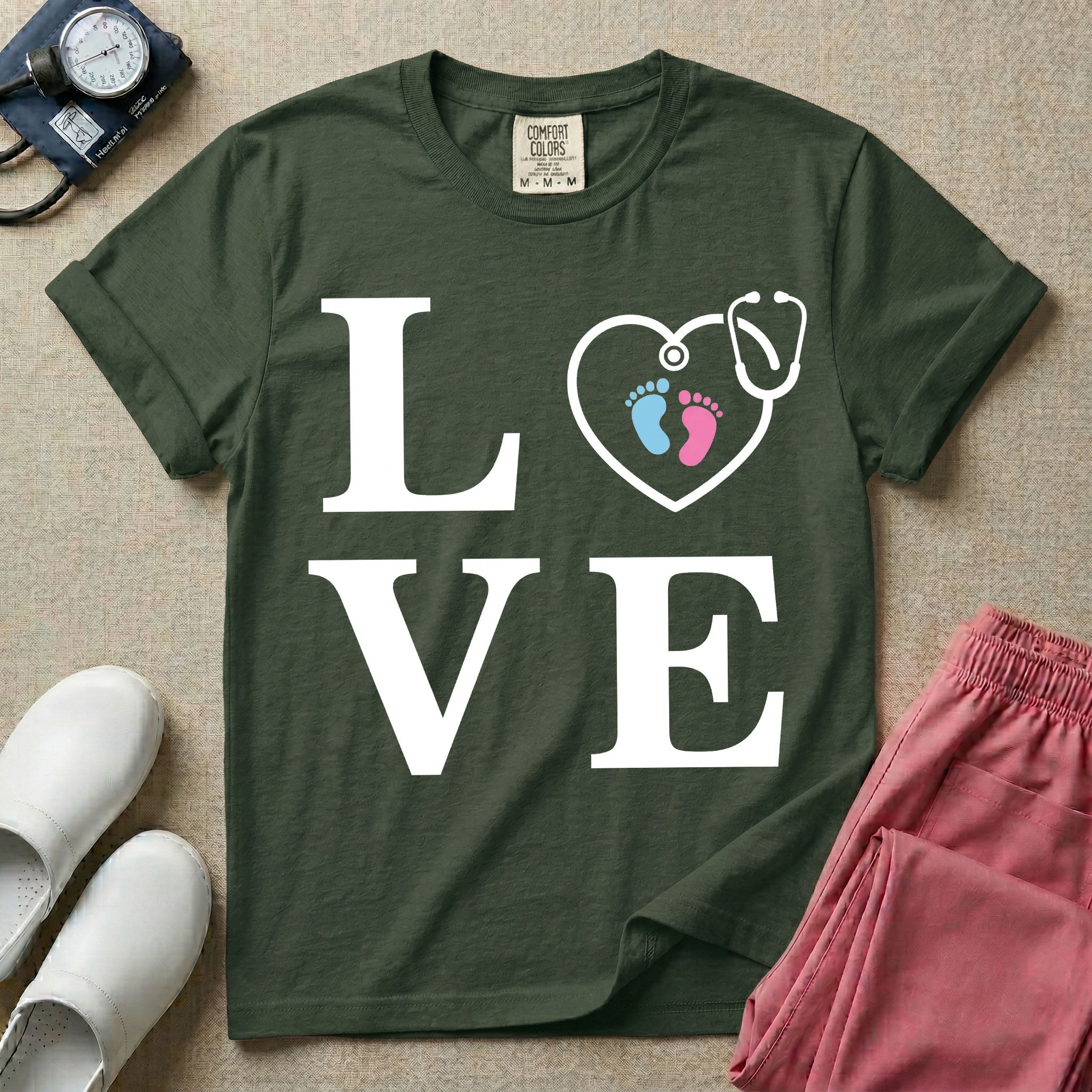 A minimalist nursing t-shirt featuring the word LOVE stacked in large white serif letters, with the letter O replaced by a white stethoscope shaped like a heart containing two small baby footprints, one pink and one blue.