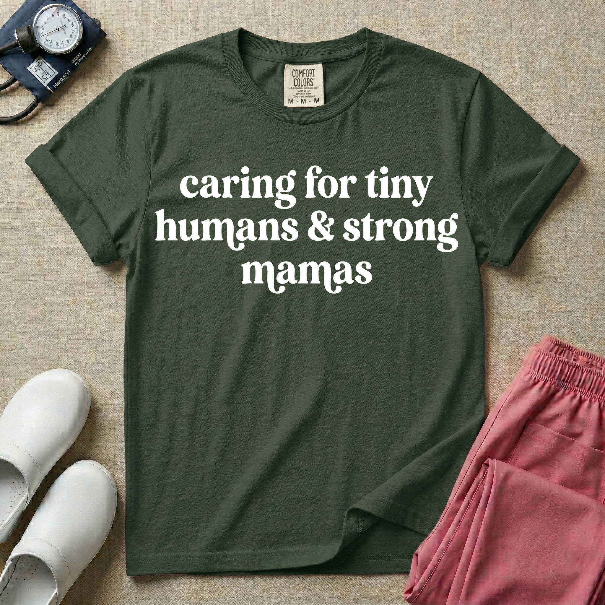 A minimalist nursing t-shirt featuring the text "caring for tiny humans & strong mamas" written in a retro white serif font.