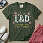 A descriptive graphic tee for labor and delivery nurses featuring plaid block letters spelling L&D surrounded by floral elements and a coffee cup, with the text "Blessed by the Babies, Stressed by the Fluids."