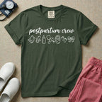 A t-shirt featuring the lowercase text postpartum crew in a cursive script font above a horizontal row of minimalist line-art icons including a baby chick, a pacifier, a baby bottle, a heart-shaped stethoscope, a swaddled baby, baby footprints, and a ribbon bow.