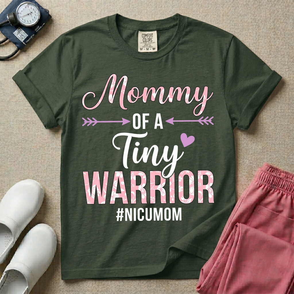 A cute NICU mom t-shirt featuring the stacked text "Mommy OF A Tiny WARRIOR #NICUMOM" in pink and white, accented with decorative arrows, a small pink heart, and a pink plaid pattern filling the word warrior.