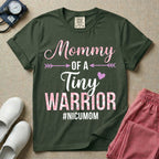 A cute NICU mom t-shirt featuring the stacked text "Mommy OF A Tiny WARRIOR #NICUMOM" in pink and white, accented with decorative arrows, a small pink heart, and a pink plaid pattern filling the word warrior.