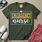 A retro-style t-shirt for emergency room nurses featuring the word "EMERGENCY" in colorful, vintage lettering above the word "nurse" in a delicate white script font, framed by a thin white rectangular border with a small heart at the top center.