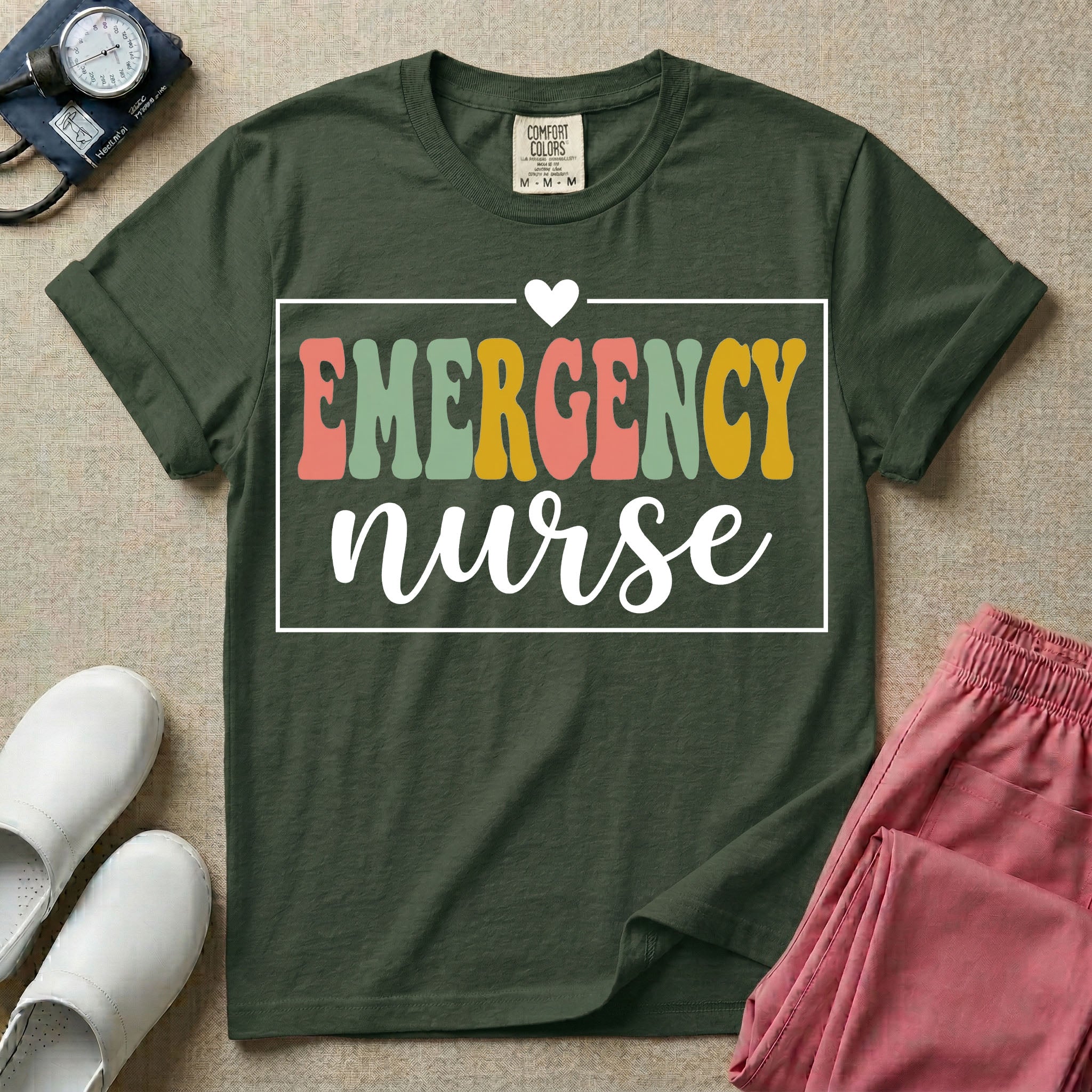 A retro-style t-shirt for emergency room nurses featuring the word "EMERGENCY" in colorful, vintage lettering above the word "nurse" in a delicate white script font, framed by a thin white rectangular border with a small heart at the top center.