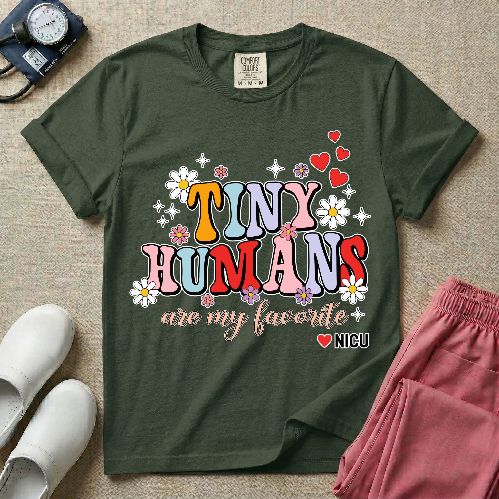 A t-shirt featuring the text Tiny Humans are my favorite in colorful retro bubble letters decorated with daisies, sparkles, and red hearts with a small NICU heart logo at the bottom.