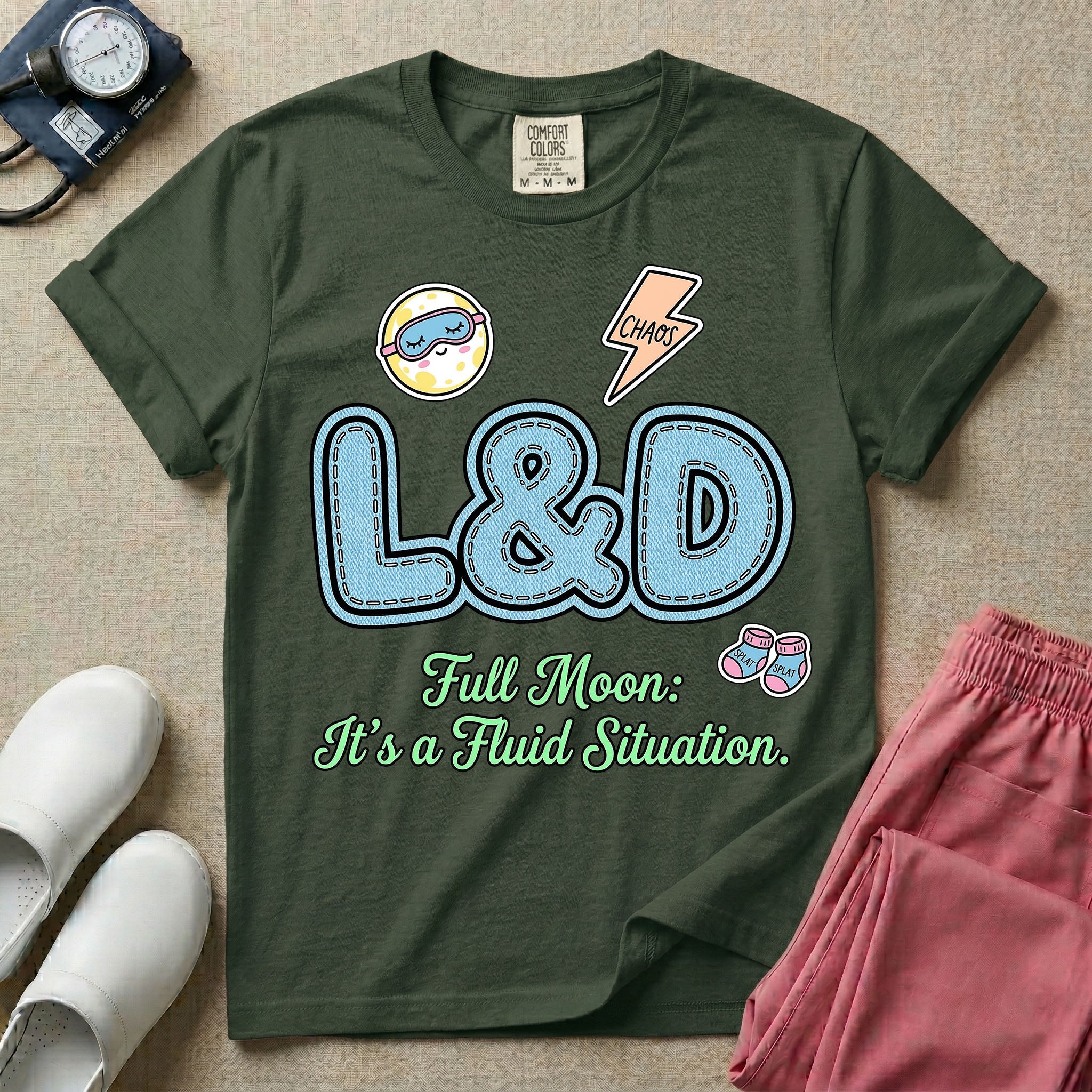 A humorous Labor and Delivery nurse graphic featuring large, light blue varsity-style letters spelling L&D with a pink dashed outline, surrounded by small patch-like icons of a baby and a lightning bolt, sitting above cursive text that reads Full Moon: It's a Fluid Situation.