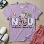 A funny t-shirt for NICU nurses featuring the large text "NICU" patterned with purple stripes and pink leopard print containing grumpy cat faces, accompanied by graphics of an IV bag with a pink bow, a steaming coffee cup reading "NICU FUEL", a green flag saying "Code Grumpy", and the bottom text "Small Babies. Shorter Fuses."