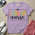 A retro-style t-shirt for emergency room nurses featuring the word "EMERGENCY" in colorful, vintage lettering above the word "nurse" in a delicate white script font, framed by a thin white rectangular border with a small heart at the top center.
