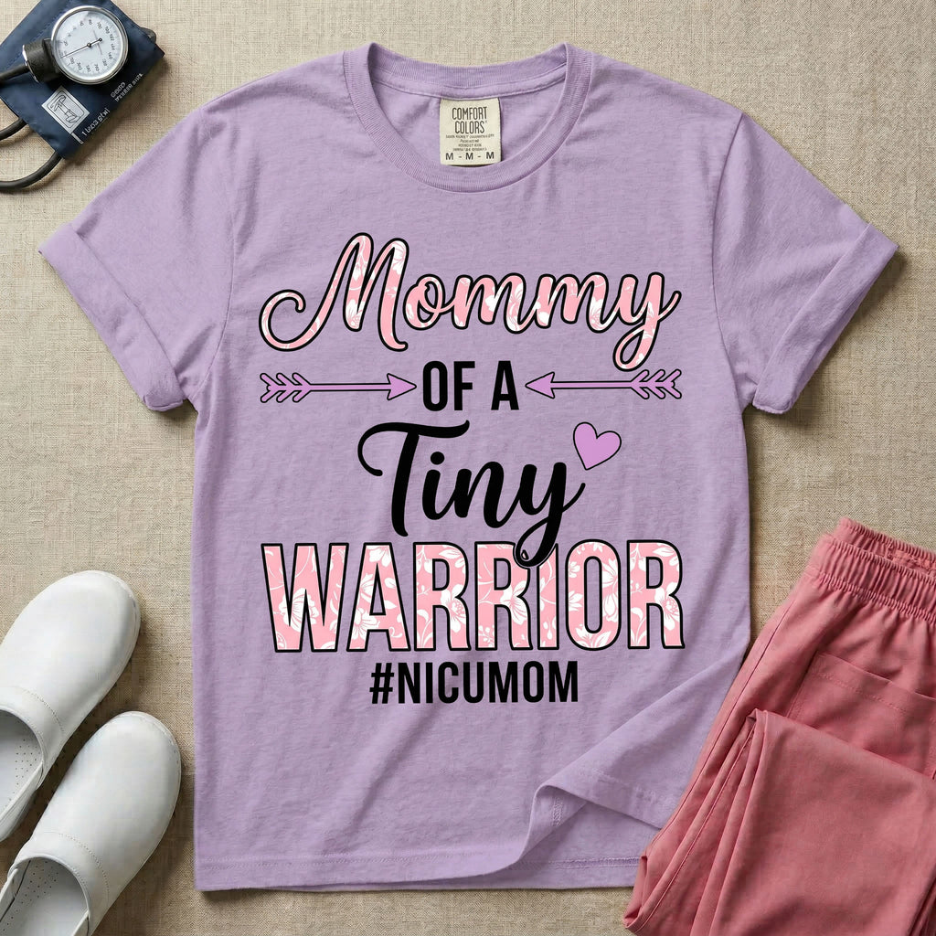 A cute NICU mom t-shirt featuring the stacked text "Mommy OF A Tiny WARRIOR #NICUMOM" in pink and white, accented with decorative arrows, a small pink heart, and a pink plaid pattern filling the word warrior.