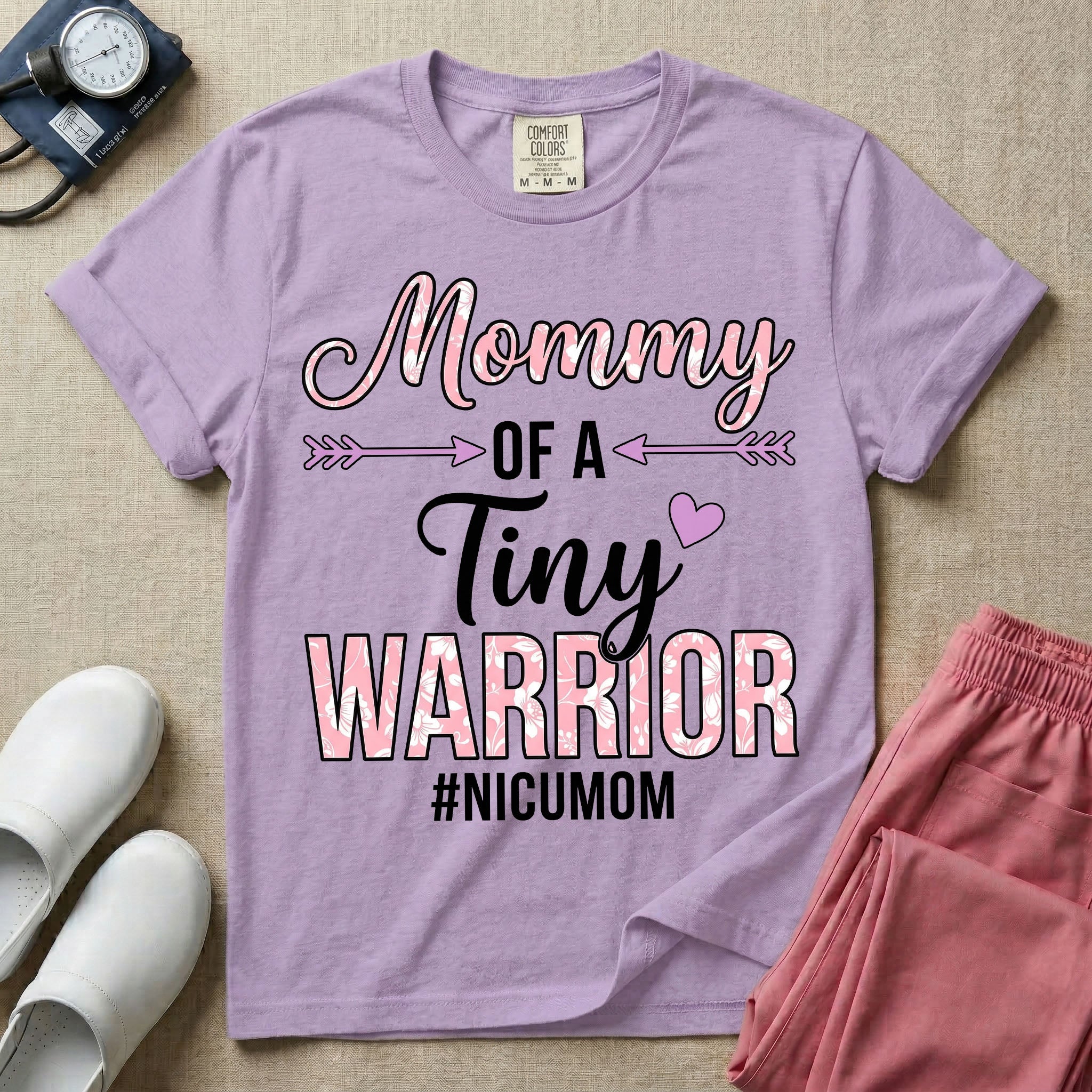 A cute NICU mom t-shirt featuring the stacked text "Mommy OF A Tiny WARRIOR #NICUMOM" in pink and white, accented with decorative arrows, a small pink heart, and a pink plaid pattern filling the word warrior.