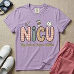 A humorous t-shirt for NICU nurses featuring the large text "NICU" in plaid and patterned letters with the phrase "Tiny Humans. Massive Attitudes." underneath, accompanied by small cute graphics of a syringe, an iced coffee cup, and a baby's face.