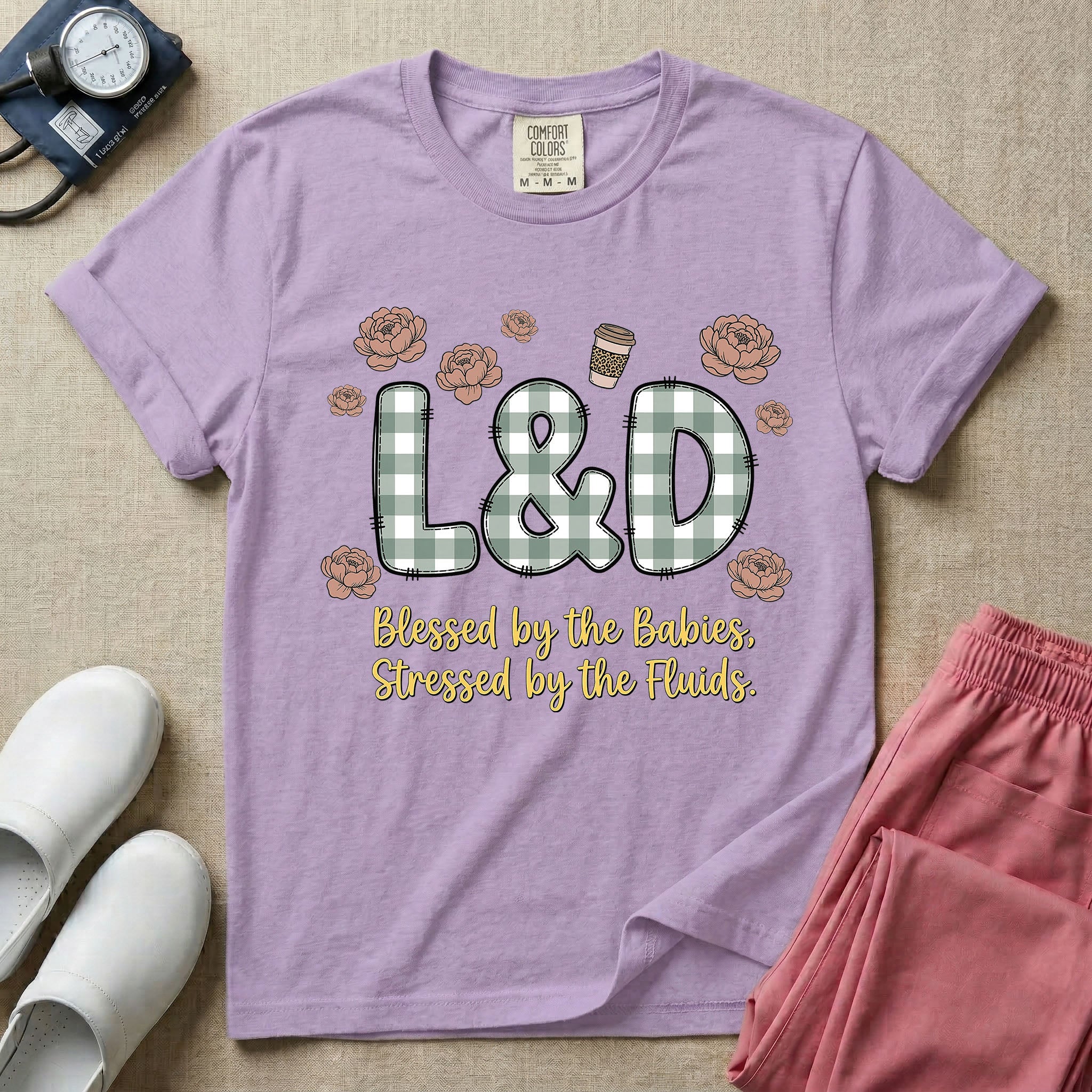 A descriptive graphic tee for labor and delivery nurses featuring plaid block letters spelling L&D surrounded by floral elements and a coffee cup, with the text "Blessed by the Babies, Stressed by the Fluids."