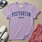 A t-shirt featuring the word POSTPARTUM arched in large collegiate block letters above the word NURSE in a smaller, matching font.