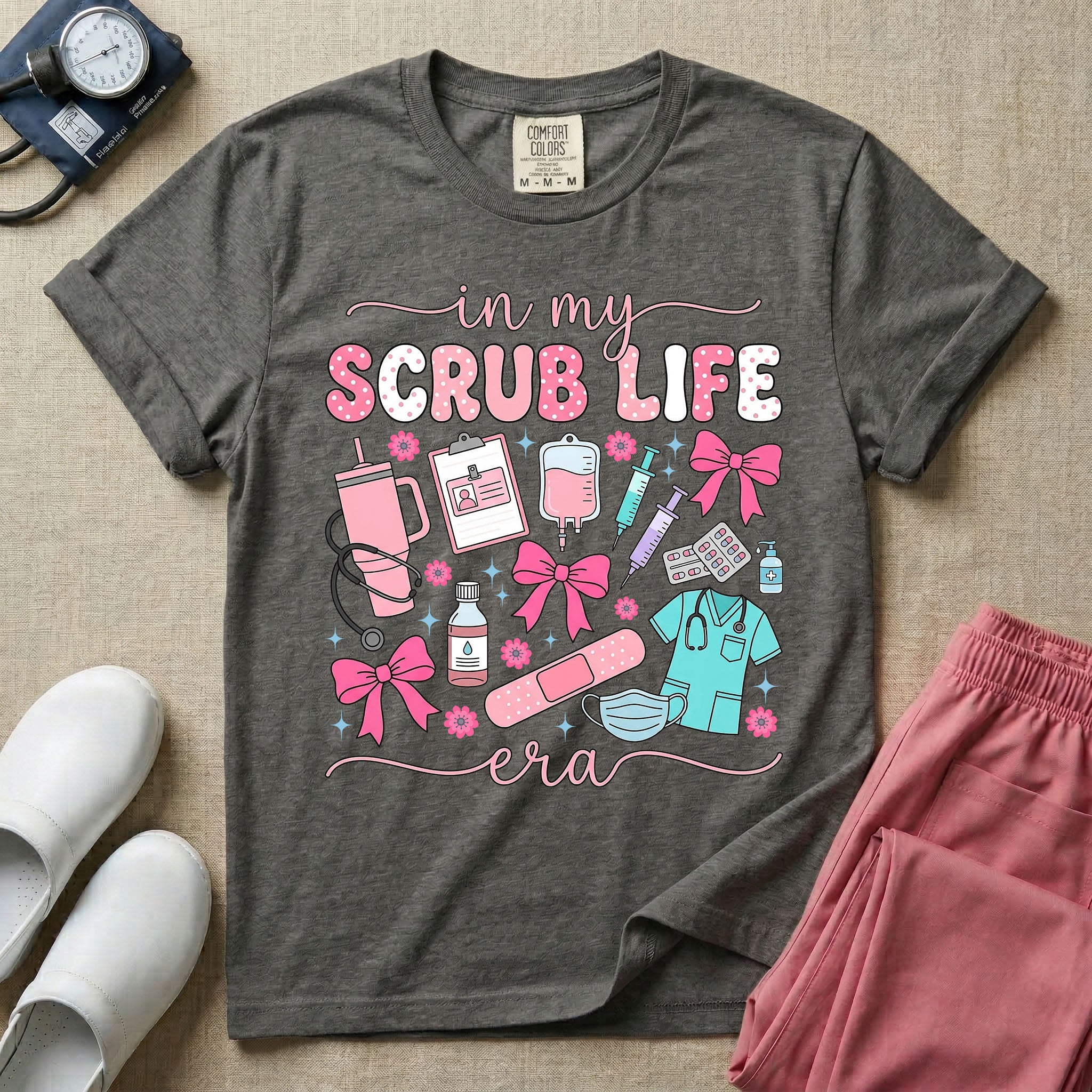 A cute general nursing t-shirt featuring the retro text "in my SCRUB LIFE era" surrounded by coquette medical graphics including a pink tumbler cup, stethoscope, IV bag with pink bows, clipboard, and blue scrubs.
