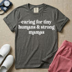 A minimalist nursing t-shirt featuring the text "caring for tiny humans & strong mamas" written in a retro white serif font.