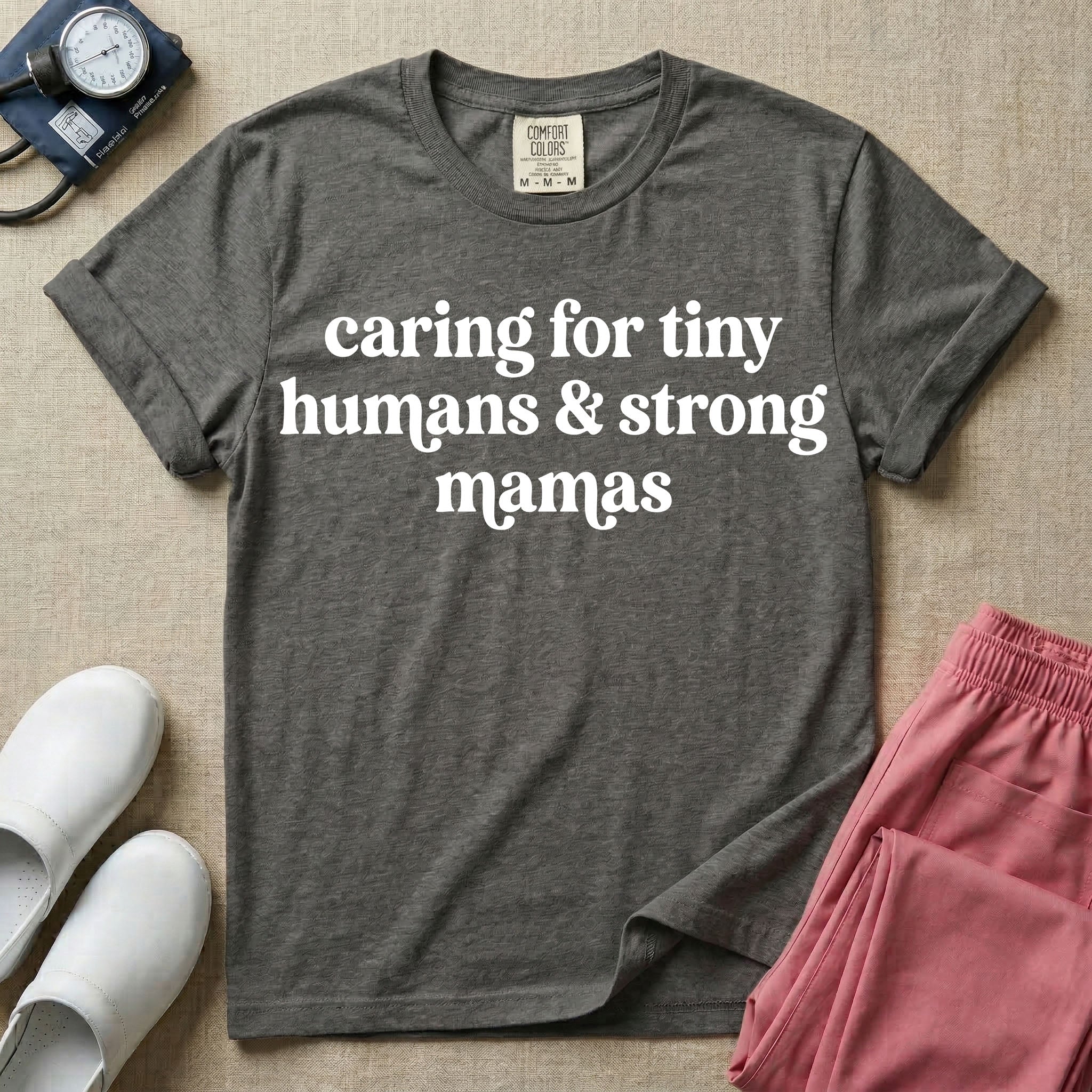 A minimalist nursing t-shirt featuring the text "caring for tiny humans & strong mamas" written in a retro white serif font.
