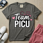 A descriptive graphic tee for pediatric intensive care nurses featuring the text "Team PICU" in bold white lettering, accented with pink hearts and a pink stethoscope design.